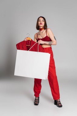 Tüketim, kısa saçlı ve burun piercingli genç bir kadın ceket askısı ve gri arka planda alışveriş çantası, modern moda eğilimi, şık tarzı, boyun atkısı, tam boy. 