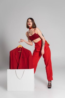 Tüketim, kısa saçlı ve burnu piercingli, spor ceketli bir kadın gri arka planda alışveriş çantasından çıkartıyor, modern moda modası, boyun atkısı, tam boy. 