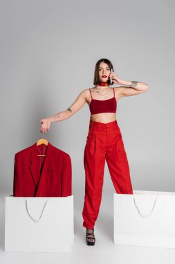 Tüketim, dövmeli kadın esmer, kısa saçlı, burun piercingli spor ceketli, akıllı telefondan konuşan gri arka planda alışveriş çantaları olan, modern moda eğilimli, uzun boylu. 
