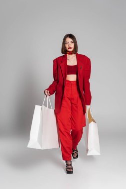 Tüketim, esmer kısa saçlı ve burunlu genç bir kadın alışveriş torbaları tutuyor ve gri arka planda yürüyor, modern moda trendi, moda kıyafeti, gençlik kültürü, tam boy 