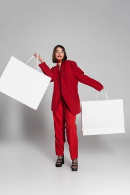 Tüketim, esmer, kısa saçlı, burun delici, alışveriş torbaları tutan, gri arka planda yürüyen, modern moda trendi, moda kıyafeti, Z jenerasyonu, tam boy. 