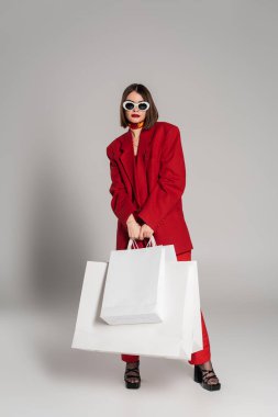 Moda, Z nesli, esmer kısa saçlı ve burun piercingli güneş gözlüklü genç bir kadın ve gri arka planda alışveriş torbaları, gençlik kültürü, kırmızı takım elbise, tüketimcilik. 