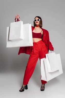Z nesli dövmeli, kısa saçlı ve burnu piercingli güneş gözlüğü ve kırmızı elbiseyle poz veren genç bir kadın. Elinde gri arka planda alışveriş torbaları, modern moda, tüketimcilik, tam boy.  