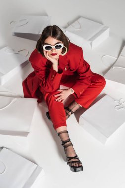 consumerism, young woman with brunette short hair, nose piercing and tattoo posing in sunglasses and red suit while sitting around shopping bags on grey background, high angle view  clipart