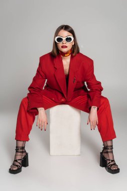 generation z, fashion model with brunette short hair and nose piercing posing in sunglasses and red suit while sitting on concrete cube on grey background, lady in red, fashionista  clipart