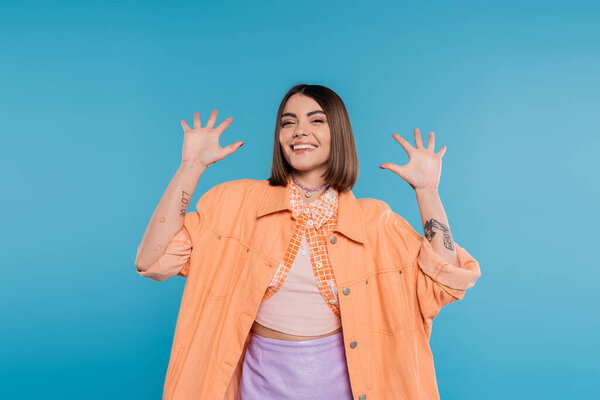 excited young woman gesturing and smiling while looking at camera, on blue background, summer outfit, generation z, short brunette hair, orange shirt, pierced nose, tattooed 