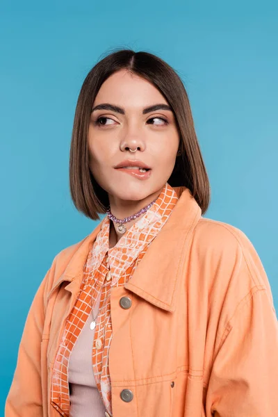 女性の肖像画彼女の唇をかむ 青の背景を離れて見て 可愛い オレンジのシャツ 世代Z 短いブルネットの髪 ピアス鼻 ジェンZファッション カジュアルな服装 — ストック写真