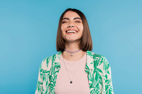 joyful young woman with short brunette hair wearing shirt with palm tree print, smiling with closed eyes on blue background, casual attire, gen z fashion, emotional, happiness, nose piercing 