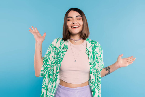 tattooed young woman with short brunette hair wearing shirt with palm tree print, smiling with closed eyes and gesturing with hands on blue background, casual attire, gen z fashion, happiness 