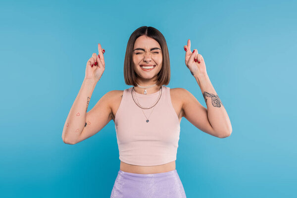 fingers crossed, tattooed young woman with short brunette hair in tank top making wish on blue background, casual attire, gen z fashion, happiness, smiling with closed eyes 