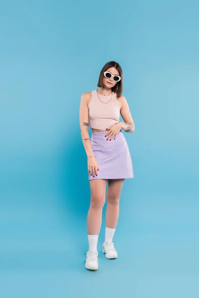 stock image fashion trend, brunette young woman with short hair in tank top, skirt, white sneakers and sunglasses posing on blue background, casual attire, gen z fashion, personal style, woman with tattoos  
