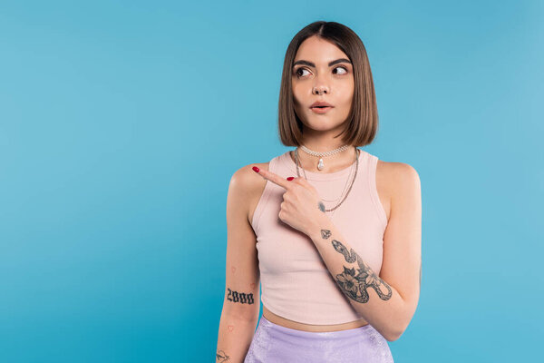 surprised woman showing something and looking away, stylish and tattooed model with nose piercing pointing with finger on blue background, generation z, emotional 