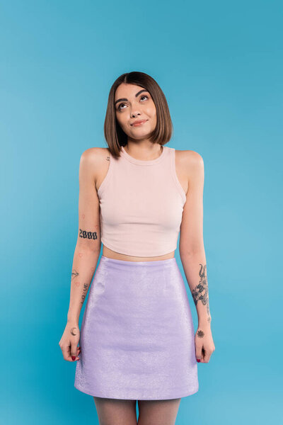 not knowing, smiling young woman with tattoos and nose piercing standing in tank top and skirt on blue background, looking up, confused, pretty face, generation z, summer outfit 