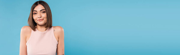 Не зная, позитивная молодая женщина с татуировками и пирсингом носа стоя в майке на синем фоне, глядя на камеру, смущенное, красивое лицо, поколение z, летний наряд, баннер 