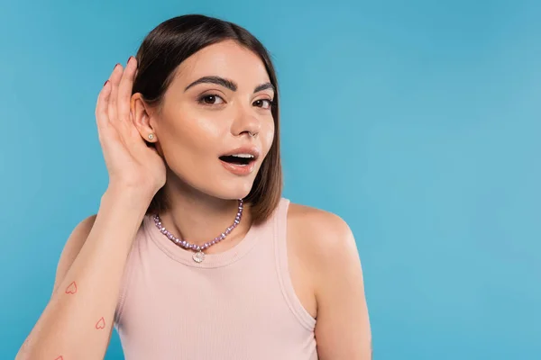 stock image Don't hear you gesture, tattooed young woman with nose piercing and short hair holding hand near ear on blue background, generation z, listening, opened mouth, curious 