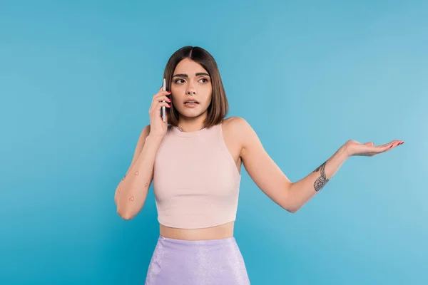 phone call, confused young woman with short hair, tattoos and nose piercing gesturing while talking on smartphone on blue background, casual attire, gen z fashion, personal style
