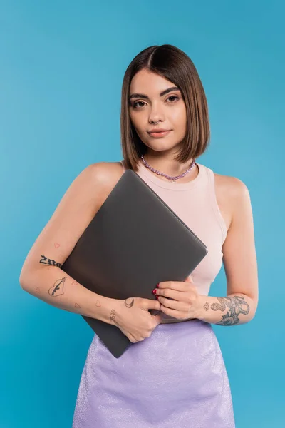 フリーランサー 短い髪を持つブルネットの若い女性 青の背景にノートパソコンを保持入れ墨や鼻ピアス 世代Z 夏の傾向 魅力的な リモートワーク 毎日のスタイル — ストック写真