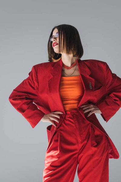 модный тренд, молодая брюнетка с короткими волосами, позирующая в розовых солнцезащитных очках и красном костюме на сером фоне, поколение Z, триумфальный наряд, модель, профессиональная экипировка 