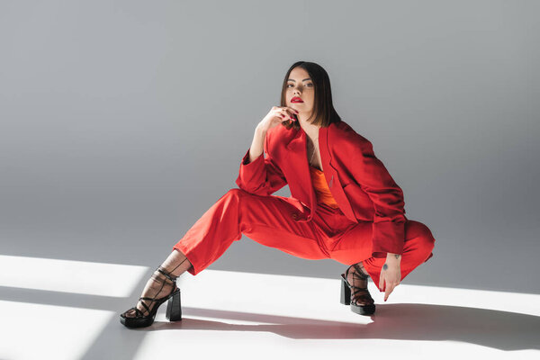 полная длина, дама в красном, молодая брюнетка женщина с короткими волосами позирует в костюме на сером фоне, поколение z, модный наряд, модная модель, профессиональный наряд, исполнительный стиль, туфли на каблуках