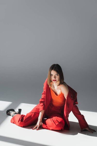 полный рост, молодая женщина с короткими волосами, сидящая в костюме на сером фоне, поколение Z, триумфальный наряд, модная модель, профессиональный наряд, стиль представительского класса, туфли на каблуках, дама в красном 