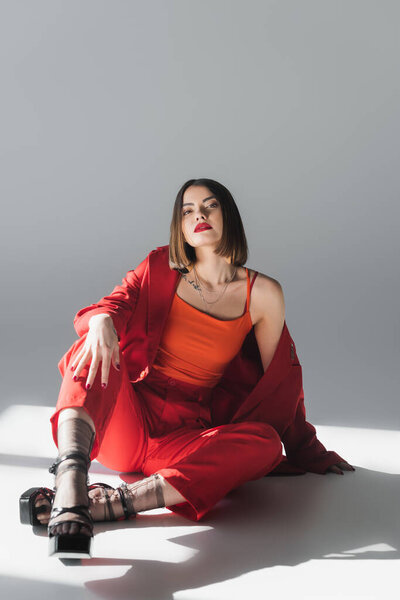 полная длина, молодая брюнетка женщина с короткими волосами сидя в костюме на сером фоне, поколение z, модный наряд, модная модель, профессиональный наряд, исполнительный стиль, туфли на каблуках, дама в красном 