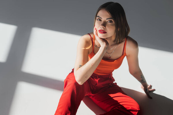 высокий угол зрения молодой татуированной женщины с короткими волосами сидя в костюме на сером фоне, поколение z, модная модель, профессиональный наряд, корпоративная мода, дама в красном 