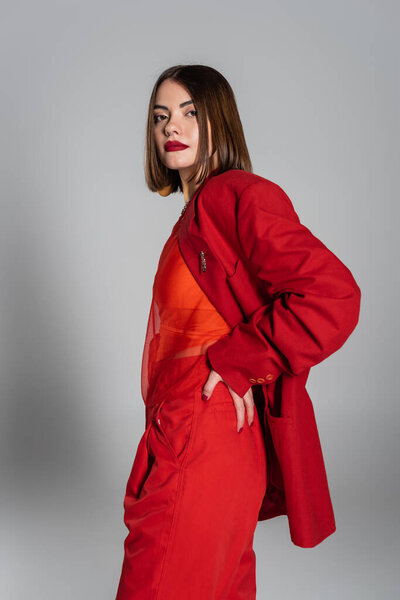 корпоративная мода, смелый макияж, молодая женщина с короткими волосами и пирсингом носа позирует в костюме на сером фоне, поколение z, модный наряд, модная модель, профессиональный наряд, рука об руку на бедре 