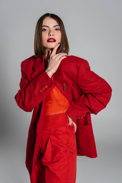 модная внешность, молодая женщина с короткими волосами и блестками под нос, позирующая в костюме на сером фоне, поколение Z, триумфальный наряд, модная модель, профессиональная экипировка, руки на бедре, смелый макияж 