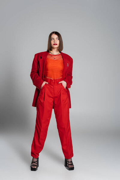 исполнительный стиль, молодая брюнетка женщина с короткими волосами и пирсинг носа позируя в костюме на сером фоне, поколение z, модный наряд, модная модель, профессиональный наряд, руки в карманах