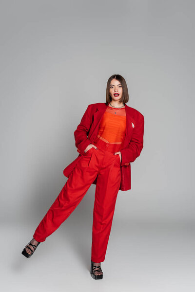 официальный наряд, молодая брюнетка женщина с короткими волосами и пирсинг носа позируя в костюме на сером фоне, поколение z, модный наряд, модная модель, профессиональный наряд, руки в карманах