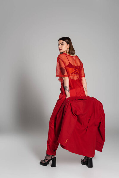 корпоративная мода, молодая брюнетка с короткими волосами позируя в костюме и держа блейзер на сером фоне, поколение z, модный наряд, модная модель, профессиональный наряд 