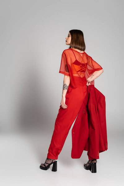 современная деловая женщина, молодая брюнетка с короткими волосами и татуировками позируя в костюме и держа блейзер на сером фоне, поколение z, модный наряд, модная модель, профессиональный наряд 