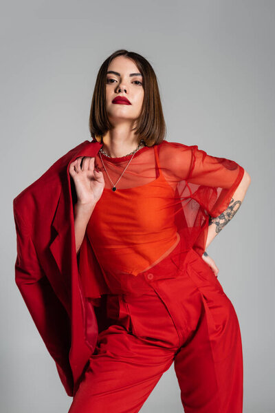 смелый макияж, исполнительный стиль, молодая татуированная женщина с короткими волосами держа красный блейзер на сером фоне, поколение z, модный наряд, модная модель, профессиональный наряд 