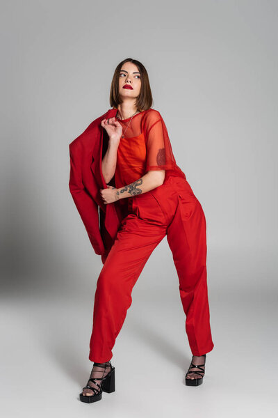 модная модель, смелый макияж, молодая татуированная женщина с короткими волосами держа красный блейзер на сером фоне, поколение z, модный наряд, профессиональный наряд, исполнительный стиль 