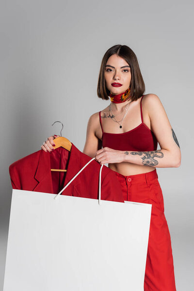 consumerism, tattooed woman with short hair and nose piercing holding hanger with blazer and shopping bag on grey background, modern fashion trend, chic style, neck scarf 