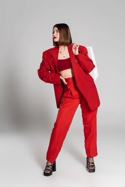 赤いスーツ 大胆なメイク ブルネットの短い髪と鼻ピアスショッピングバッグを保持し 灰色の背景 若者文化 ファッショナブルな衣装 消費者主義に立つ若い女性の入れ墨 — ストック写真