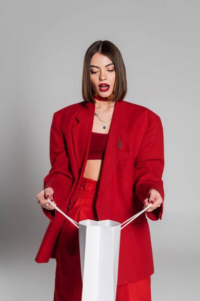 fashionable, generation z, shocked young woman with brunette short hair and nose piercing looking inside of shopping bag on grey background, youth culture, red suit, consumerism 
