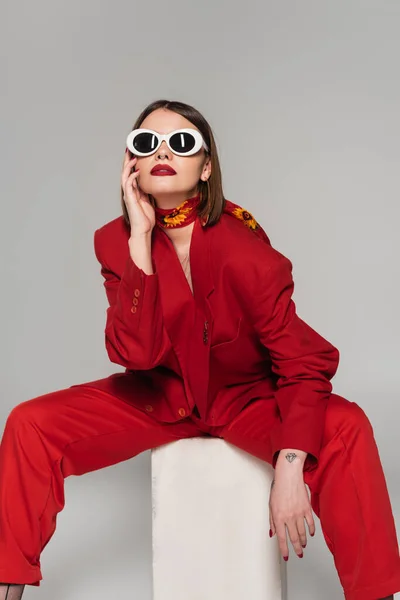 stock image generation z, fashion model with brunette short hair and nose piercing posing in sunglasses and red suit while sitting on concrete cube on grey background, lady in red, young woman in red outfit 