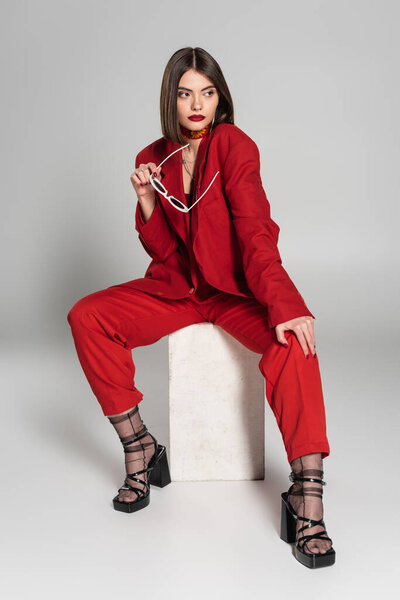 fashion model with brunette short hair and nose piercing posing in red suit while holding sunglasses and sitting on concrete cube on grey background, lady in red, young woman, fashion trend 