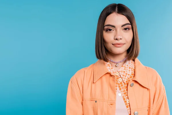 generation z, portrait of pretty woman, young fashion model looking at camera on blue background, orange shirt, short brunette hair, pierced nose, summer outfit, gen z fashion