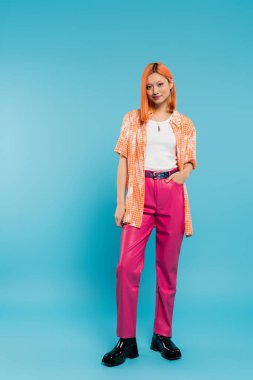 El ele, gülümseyen ve turuncu gömlekli, mavi arka planda poz veren Asyalı genç bir kadın, kameraya, pembe pantolona, Z kuşağına, modern tarzda, tam boy 