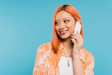 pozitiflik, telefon görüşmesi, neşeli ve genç Asyalı kadın akıllı telefondan konuşuyor ve mavi arka plandan başka tarafa bakıyor, günlük tarz, moda, sohbet, cep telefonu iletişimi 