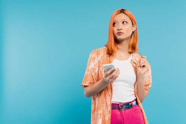Sosyal medya etkileyicisi, boyalı saçlı, akıllı telefonlu düşünceli Asyalı kadın mavi arka plan, cep telefonu, gençlik kültürü, dijital yaş, z nesline bakıyor. 