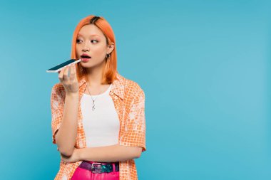 Sosyal medya etkileyicisi, boyalı saçlı genç Asyalı kadın akıllı telefon tutuyor ve mavi arka plan, cep telefonu, gençlik kültürü, dijital yaş, z nesli üzerine sesli mesaj kaydediyor. 
