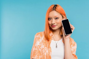 Sosyal medya etkeni, mutlu ve genç Asyalı kadın boyalı saçlı elinde mavi arka planda boş ekranlı akıllı telefon cep telefonu, cep telefonu, gençlik kültürü, dijital yaş, z nesli 