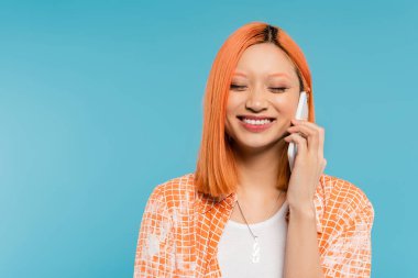 Telefon görüşmesi, pozitiflik, neşeli ve genç Asyalı saçları boyalı, gözleri kapalı mavi arka planda akıllı telefonla konuşan, cep telefonu, gençlik kültürü, dijital yaş, z nesli.