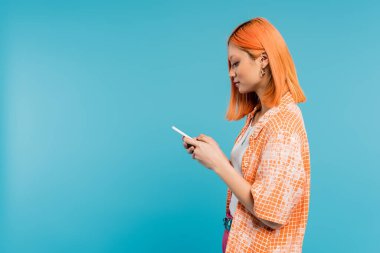 Sosyal medya etkileyicisi, boyalı saçlı genç Asyalı kadın mavi arka planda akıllı telefon kullanıyor, cep telefonu, gençlik kültürü, dijital yaş, z nesli, mesajlaşma, kenar görüş 