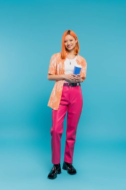 cheerful asian and young woman with red hair holding paper cup and looking at camera on blue background, casual attire, generation z, coffee culture, hot drink, single use cup, full length clipart