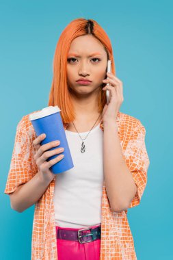 phone call, upset asian woman with red hair holding coffee to go in paper cup and talking on smartphone on blue background, casual attire, generation z, coffee culture, displeased, pouting lips  clipart