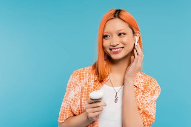 summer happiness, music lover, young and trendy asian woman with dyed hair, in orange shirt holding case, adjusting wireless earphone and looking away on blue background, generation z clipart
